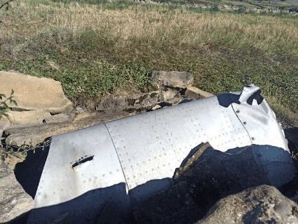 fragmento de fuselaje del ala de un Katiuska en un entorno rural y árido. La pieza, de color plateado mate, destaca por su superficie curva cubierta de hileras regulares de remaches industriales, Presenta daños por impacto, bordes deformados y una pequeña perforación en el lateral izquierdo. El resto metálico yace sobre un terreno rocoso, rodeado de piedras grandes y tierra seca. Al fondo, se aprecia una ladera con vegetación baja y pasto amarillento bajo una luz solar intensa que proyecta sombras profundas. La escena transmite una sensación de abandono histórico, donde un componente de ingeniería avanzada ha quedado integrado de forma discordante en un paisaje natural solitario.
